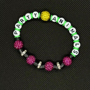 May include: A beaded bracelet with the words "PARTY ANIMALS". The bracelet features white letter beads, green and black spacers, and three fuchsia-colored rhinestone beads. A yellow rhinestone bead is also present.