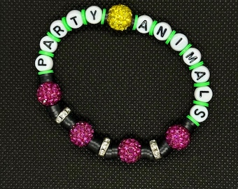 Può includere: Un braccialetto di perline con la scritta "PARTY ANIMALS". Il braccialetto presenta perline con lettere bianche, distanziatori verdi e neri e tre perline di strass fucsia. È presente anche una perlina di strass gialla.