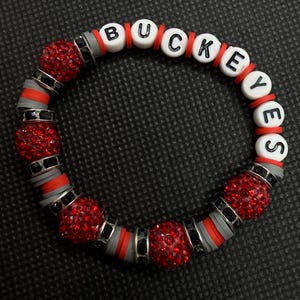 May include: A circular bracelet featuring white letter beads spelling BUCKEYES, interspersed with red glittery spherical beads, flat grey and red clay discs, and small black metal spacers. The design alternates between the lettered section and the textured beads.