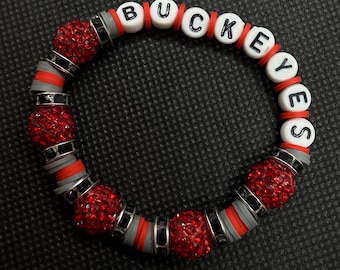 Può includere: Un braccialetto circolare con perline bianche con lettere che compongono la scritta BUCKEYES, alternate a perline sferiche rosse glitterate, dischi piatti in argilla grigia e rossa e piccoli distanziatori in metallo nero lungo tutta la circonferenza.