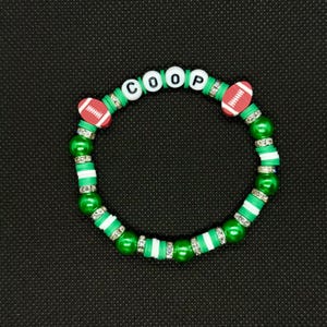 May include: A green and white beaded bracelet with the name "COOP" spelled out in white letter beads. The bracelet features two red and white football-shaped beads and is accented with silver-colored spacer beads.