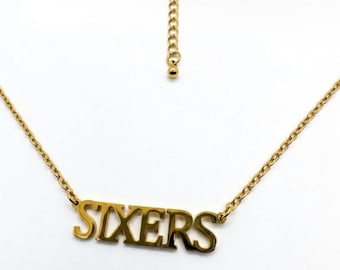 Può includere: Una collana in metallo color oro con un ciondolo che riporta la parola SIXERS in un carattere a blocchi. Il ciondolo è fissato a una delicata catena a maglie che include una chiusura a moschettone e una catenella di estensione con una piccola perlina.