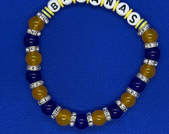 Può includere: Un braccialetto di perline con la scritta "BANANAS" in perline di lettere bianche. Il braccialetto presenta perline gialle e blu navy alternate, con distanziatori di strass argentati. Lo sfondo è blu unito.