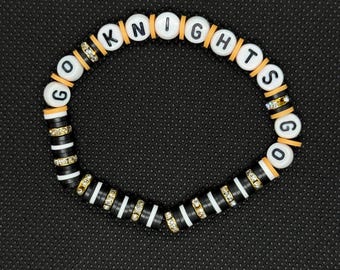 Può includere: Un braccialetto di perline con le parole "GO KNIGHTS" scritte con perline di lettere bianche. Il braccialetto presenta perline a righe bianche e nere, distanziatori arancioni e perline di strass dorate. Il braccialetto è su uno sfondo nero.