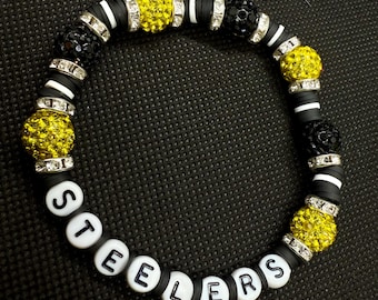 Può includere: Un braccialetto di perline bianche e nere con la parola "Steelers" scritta con perline di lettere bianche. Il braccialetto presenta perline decorative gialle e nere e dettagli argentati. Il braccialetto è su una superficie nera.