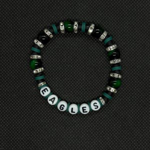 May include: A beaded bracelet with the word "EAGLES" spelled out in white letter beads. The bracelet features black, green, and silver beads. The background is a dark gray.
