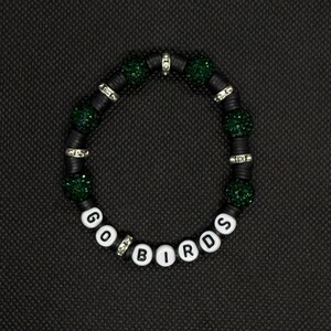 May include: A black beaded bracelet with the words "GO BIRDS" spelled out in white letter beads. The bracelet features green glittery beads and silver-toned spacers. The bracelet is displayed on a black surface.