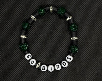 Può includere: Un braccialetto di perline nere con le parole "GO BIRDS" scritte con perline di lettere bianche. Il braccialetto presenta perline verdi glitterate e distanziatori argentati. Il braccialetto è esposto su una superficie nera.