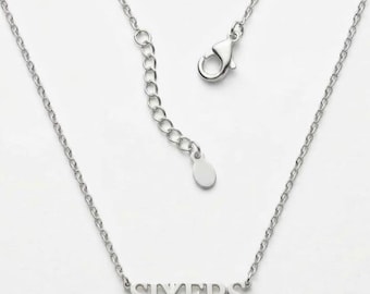 Può includere: Una collana color argento con un ciondolo centrale che riporta la parola SIXERS in carattere stampatello. La catenina include una chiusura a moschettone e una catenella di estensione regolabile con una piccola targhetta metallica ovale e liscia.