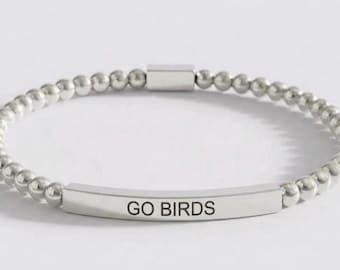 Può includere: Un braccialetto di perline color argento con una barra metallica curva centrale incisa con il testo GO BIRDS in lettere maiuscole nere. Il braccialetto è composto da un filo continuo di piccole sfere metalliche uniformi e una chiusura.