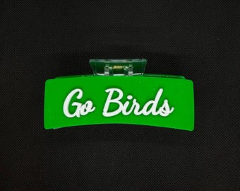 Può includere: Un fermaglio per capelli rettangolare verde brillante con la scritta bianca Go Birds in carattere corsivo. L'accessorio presenta un meccanismo a cerniera in plastica trasparente nella parte superiore centrale, progettato per fissare i capelli.