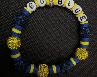 Può includere: Un braccialetto circolare con perline a lettere bianche che compongono GO BLUE, intervallate da perline a disco piatte gialle e blu, sfere gialle scintillanti incastonate di strass e sfere blu scuro testurizzate, con una perlina distanziatrice color argento.