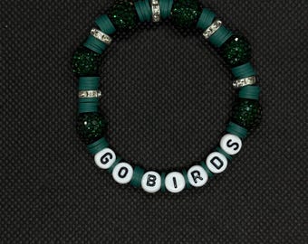 May include: A green and white beaded bracelet with the words "GO BIRDS". The bracelet features dark green beads, silver accents, and white letter beads. The bracelet is on a black background.