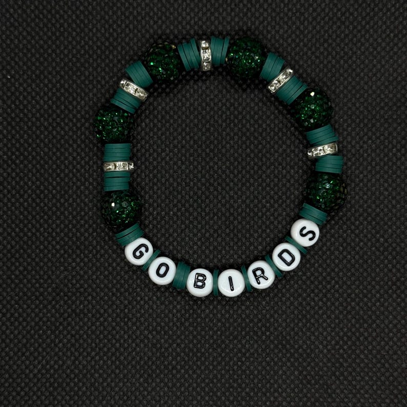 May include: A green and white beaded bracelet with the words "GO BIRDS". The bracelet features dark green beads, silver accents, and white letter beads. The bracelet is on a black background.