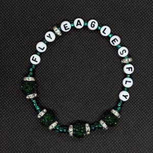 May include: A beaded bracelet with the words "FLY EAGLES FLY" spelled out in white letter beads. The bracelet features green beads and silver accents, set against a dark background. The bracelet is a fan accessory.