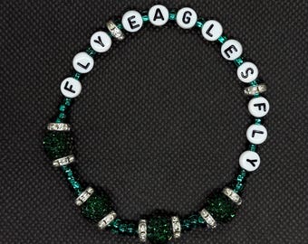 Può includere: Un braccialetto di perline con le parole "FLY EAGLES FLY" scritte con perline di lettere bianche. Il braccialetto presenta perline verdi e dettagli argentati, su uno sfondo scuro. Un accessorio per fan.