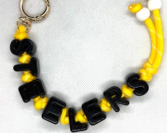 Può includere: Un portachiavi con un anello in metallo dorato attaccato a un cordino in paracord giallo. Il cordino è infilato con perline a forma di lettera in plastica nera che compongono la parola STEELERS, rifinito con due perline sferiche bianche.