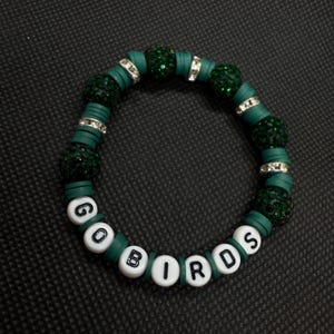 May include: A green beaded bracelet with the words "GO BIRDS" spelled out in white letter beads. The bracelet features dark green beads, flat green disc beads, and silver-colored spacer beads. The bracelet is on a black surface.