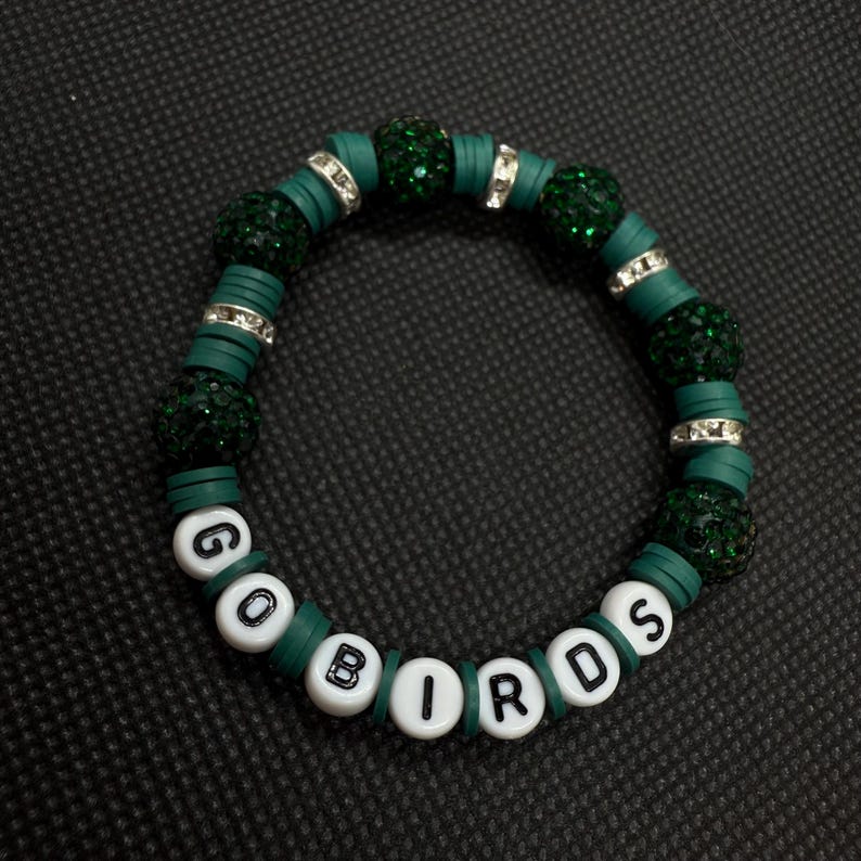 May include: A green beaded bracelet with the words "GO BIRDS" spelled out in white letter beads. The bracelet features dark green beads, flat green disc beads, and silver-colored spacer beads. The bracelet is on a black surface.