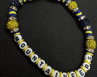 Può includere: Un braccialetto di perline con dischi piatti gialli e blu navy, intervallati da sfere scintillanti incastonate di strass gialli e blu scuro. Perline bianche con lettere nere compongono la parola MOUNTAINEERS in maiuscolo.