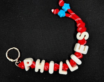 Può includere: Un portachiavi con un cordino rosso a macchie bianche, annodato tra perline bianche con lettere che compongono la parola PHILLIES. Il cordino termina con due perline a forma di stella blu brillante e un moschettone in metallo dorato.