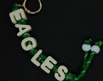 Può includere: Un portachiavi con un anello in metallo dorato attaccato a un cordino verde e bianco maculato. Il cordino è infilato con perline a lettera in plastica color avorio che compongono la parola EAGLES, rifinito con due perline sferiche bianche.