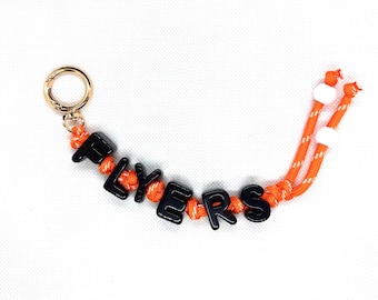 Può includere: Un portachiavi in paracord arancione con filo riflettente bianco, perline con lettere nere che compongono la parola FLYERS e un moschettone in metallo dorato. Due perline bianche sono fissate alle estremità del cordino arancione annodato.