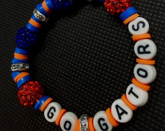 Può includere: Un braccialetto di perline con dischi piatti blu e arancioni, intervallati da distanziatori argentati con strass e perline sferiche testurizzate rosse e blu. Perline bianche con lettere nere compongono la scritta GO GATORS.