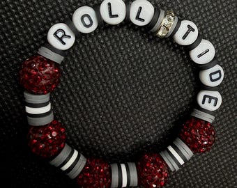 Può includere: Un braccialetto circolare di perline con perline rotonde bianche con lettere nere che compongono ROLL TIDE, intervallate da perline sferiche rosso scuro glitterate, perline a disco piatte grigie, strisce bianche e una singola perlina distanziatrice in strass argentato.