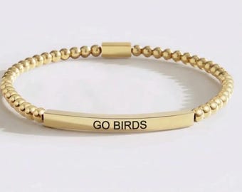 Può includere: Un braccialetto di perline color oro con una barra metallica curva centrale incisa con il testo GO BIRDS in lettere maiuscole nere. Il braccialetto è composto da una serie continua di piccole perline metalliche uniformi e una chiusura.