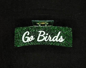 Può includere: Un fermaglio per capelli rettangolare con una finitura glitterata verde brillante. Il testo Go Birds è visualizzato al centro in un carattere corsivo bianco e grassetto. La clip include un meccanismo a cerniera in plastica trasparente con una molla dorata.
