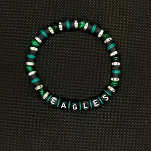 May include: A beaded bracelet with the word "EAGLES" spelled out in white letter beads. The bracelet features black, green, and silver beads. The background is a dark, textured surface.
