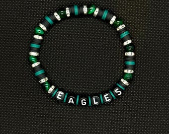 Può includere: Un braccialetto di perline con la parola "EAGLES" scritta con perline di lettere bianche. Il braccialetto presenta perline nere, verdi e argentate. Lo sfondo è una superficie scura e strutturata.