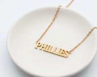 Può includere: Una collana in metallo color oro con un ciondolo a targhetta che riporta la parola PHILLIES in un carattere serif maiuscolo e in grassetto. Il ciondolo è fissato a una sottile e delicata catenina color oro che si estende verso l'alto.