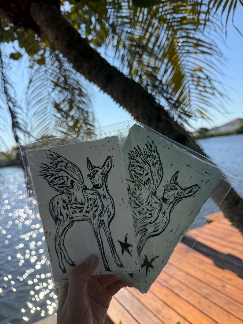 K&ouml;nnte beinhalten: Zwei Linolschnitt-Drucke mit einem gefl&uuml;gelten Hirsch und einem Stern. Ein Druck ist auf wei&szlig;em Papier, der andere auf hellgr&uuml;nem Papier. Die Drucke befinden sich in transparenten Kunststoffh&uuml;llen, vor dem Hintergrund von Wasser und einem Holzsteg.