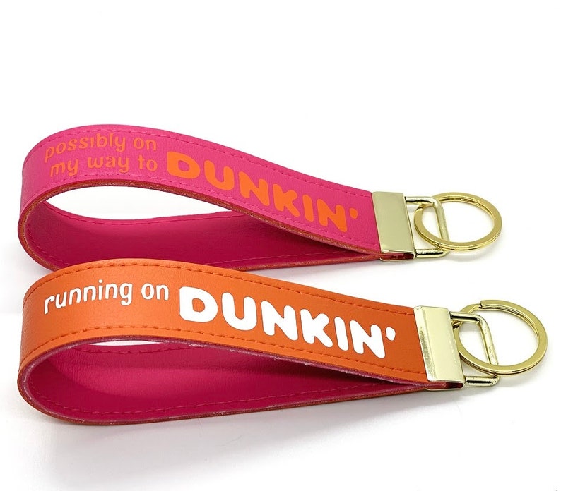 May include: Two pink and orange leather keychains with gold key rings. The keychains have the text "possibly on my way to DUNKIN'" and "running on DUNKIN'" printed on them.