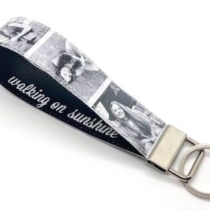 May include: Black and white photo keychain with a black leather strap. The strap has the text "walking on sunshine" printed on it. The keychain has a silver metal ring.