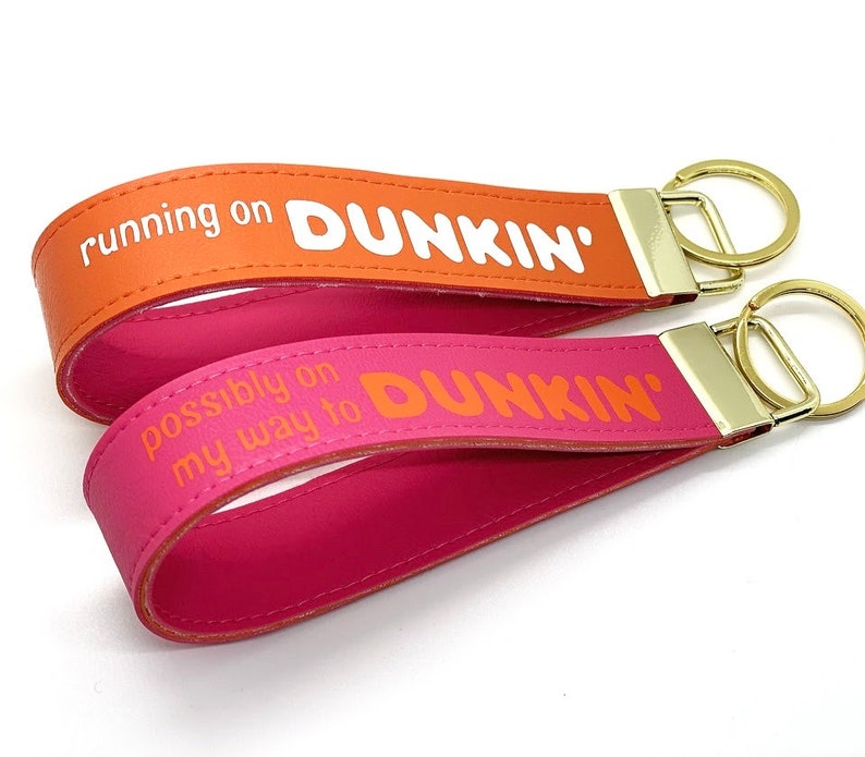 May include: Two leather keychains with gold metal key rings. The top keychain is orange with the text "running on DUNKIN'" and the bottom keychain is pink with the text "possibly on my way to DUNKIN'".