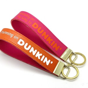 May include: Two pink and orange leather keyrings with gold key rings. The keychains have the text "running on Dunkin'" and "possibly on my way to Dunkin'" printed on them.
