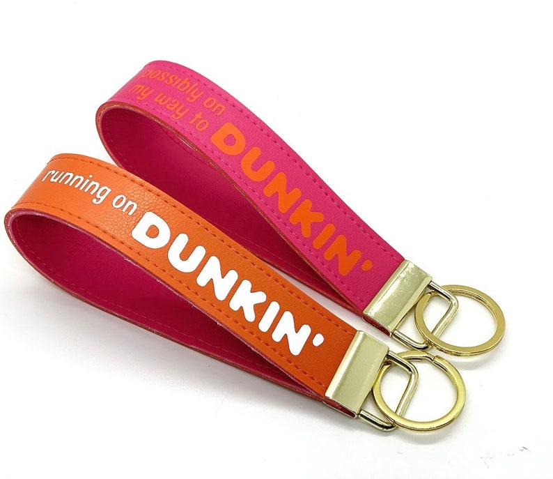 May include: Two pink and orange leather keyrings with gold key rings. The keychains have the text "running on Dunkin'" and "possibly on my way to Dunkin'" printed on them.