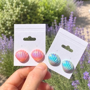 May include: Two pairs of shell-shaped stud earrings attached to white display cards. One pair features a glossy, iridescent peach-pink finish, while the other pair displays a shimmering, iridescent blue-green finish with a radial ribbed texture.