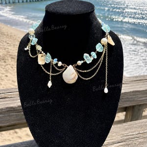 May include: A multi-layered necklace featuring light blue translucent stone chips, white pearls, and small gold-toned metal chains. A central teardrop-shaped shell pendant hangs from the gold chain, accented by additional small shell charms.