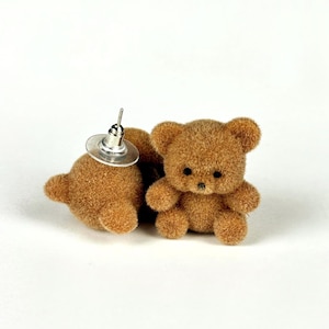 May include: A pair of brown teddy bear stud earrings. Each earring features a small, plush teddy bear with black bead eyes and a silver post. The earrings are set against a white background.