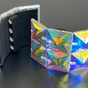 Pode incluir: Uma pulseira segmentada com cristais iridescentes de corte quadrado. A pulseira é composta por várias secções articuladas, permitindo flexibilidade. O fecho é preto e a moldura é transparente. Os cristais refletem um espectro de cores, criando um efeito deslumbrante.