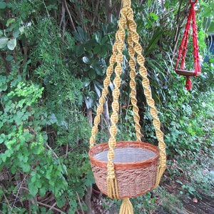 May include: A woven brown basket plant holder suspended by a knotted, golden-yellow macrame hanger. The basket has a clear plastic liner. A red macrame plant holder is visible in the background.