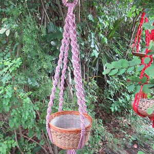 May include: A pink macrame plant hanger with a woven basket. The hanger is hanging from a tree branch in a garden setting.