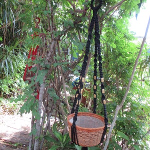 May include: A black macrame plant hanger with wooden beads hangs from a tree branch. The hanger is holding a brown woven basket planter.