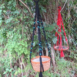 May include: Two macrame plant hangers with different colored cords and beads. The black hanger has teal beads and a brown woven pot. The red hanger has a brown woven tray.