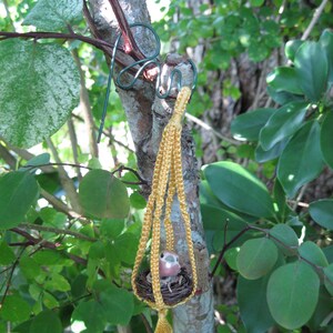 May include: A small bird figurine in a yellow macrame hanging nest, suspended from a tree branch.