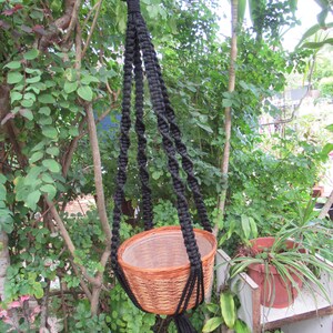 May include: A black macrame plant hanger with a woven basket pot. The plant hanger is suspended from a tree branch, showcasing a natural, bohemian aesthetic. The basket is a light brown color, and the hanger is made of thick, black rope.